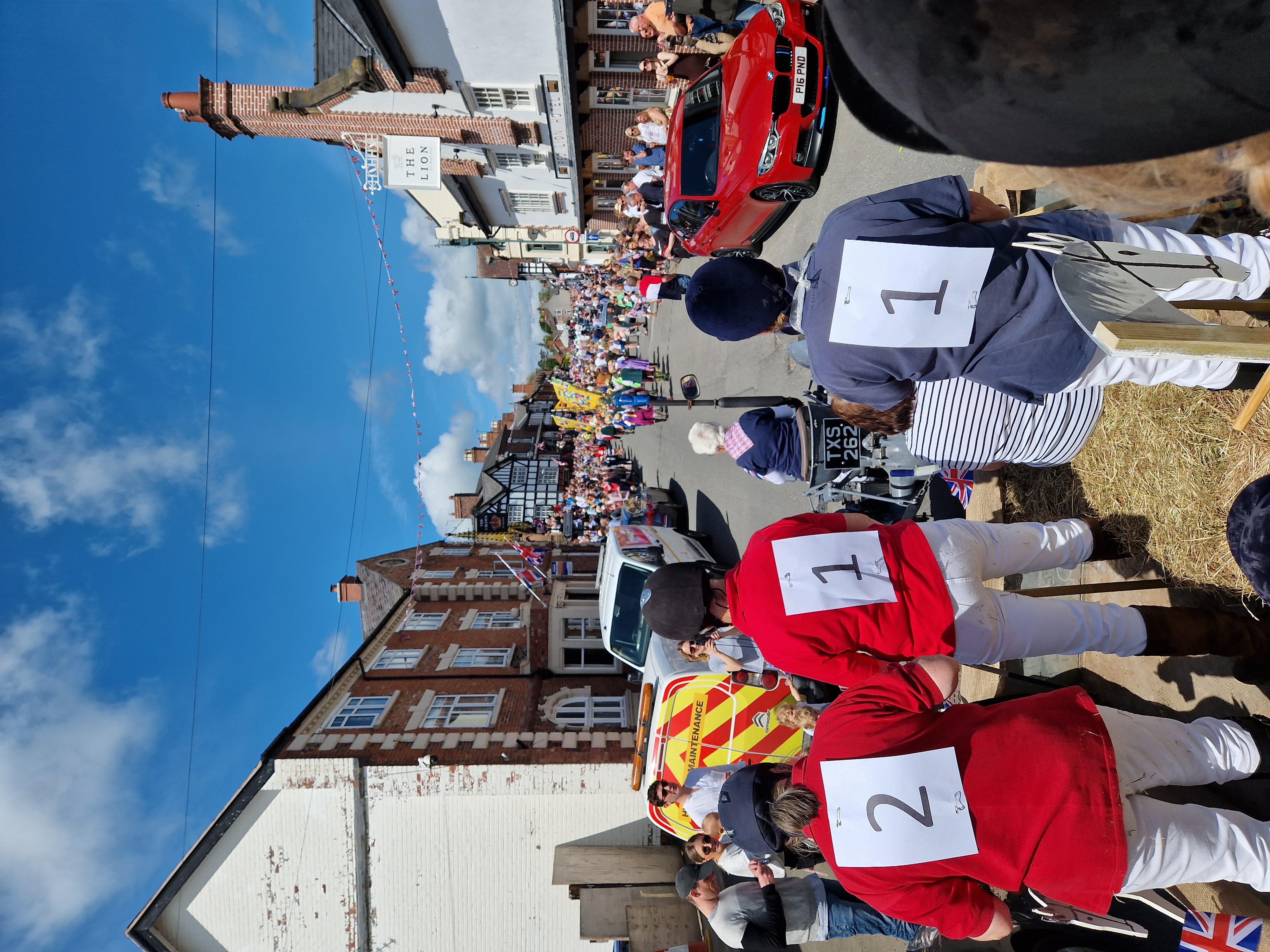 A lively village street parade with crowds lining the road on a sunny day.